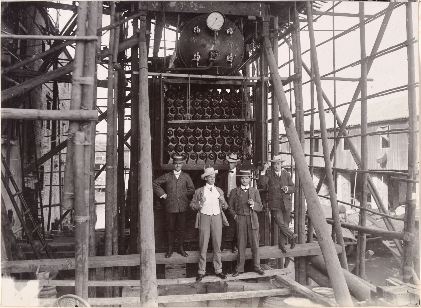 Five men in suits and boaters on a plank platform in front of a large boiler, inside bamboo scaffolding.