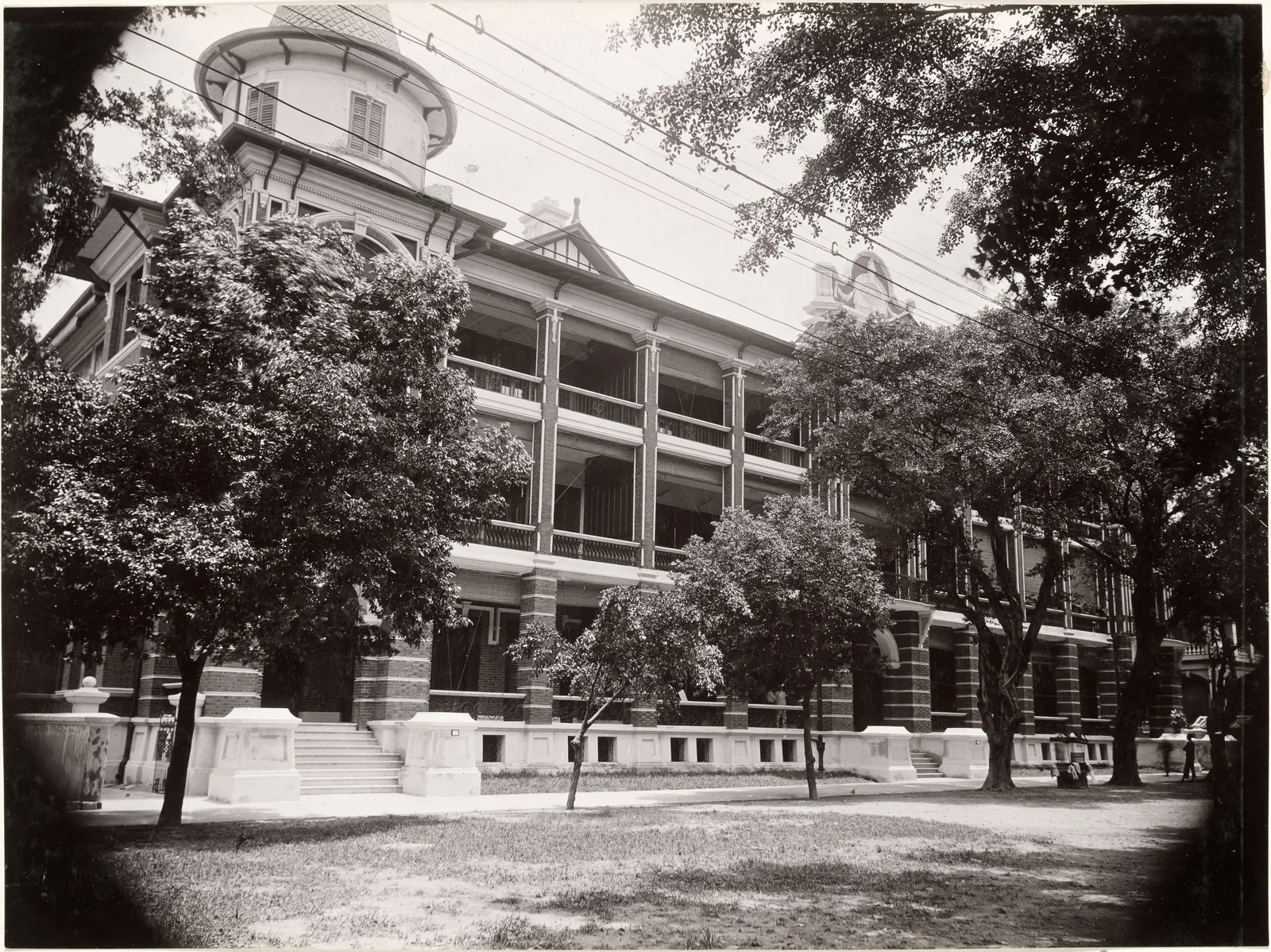 A long three-storey brick building with deep verandahs and a corner turret, seen through trees in a round vignette.