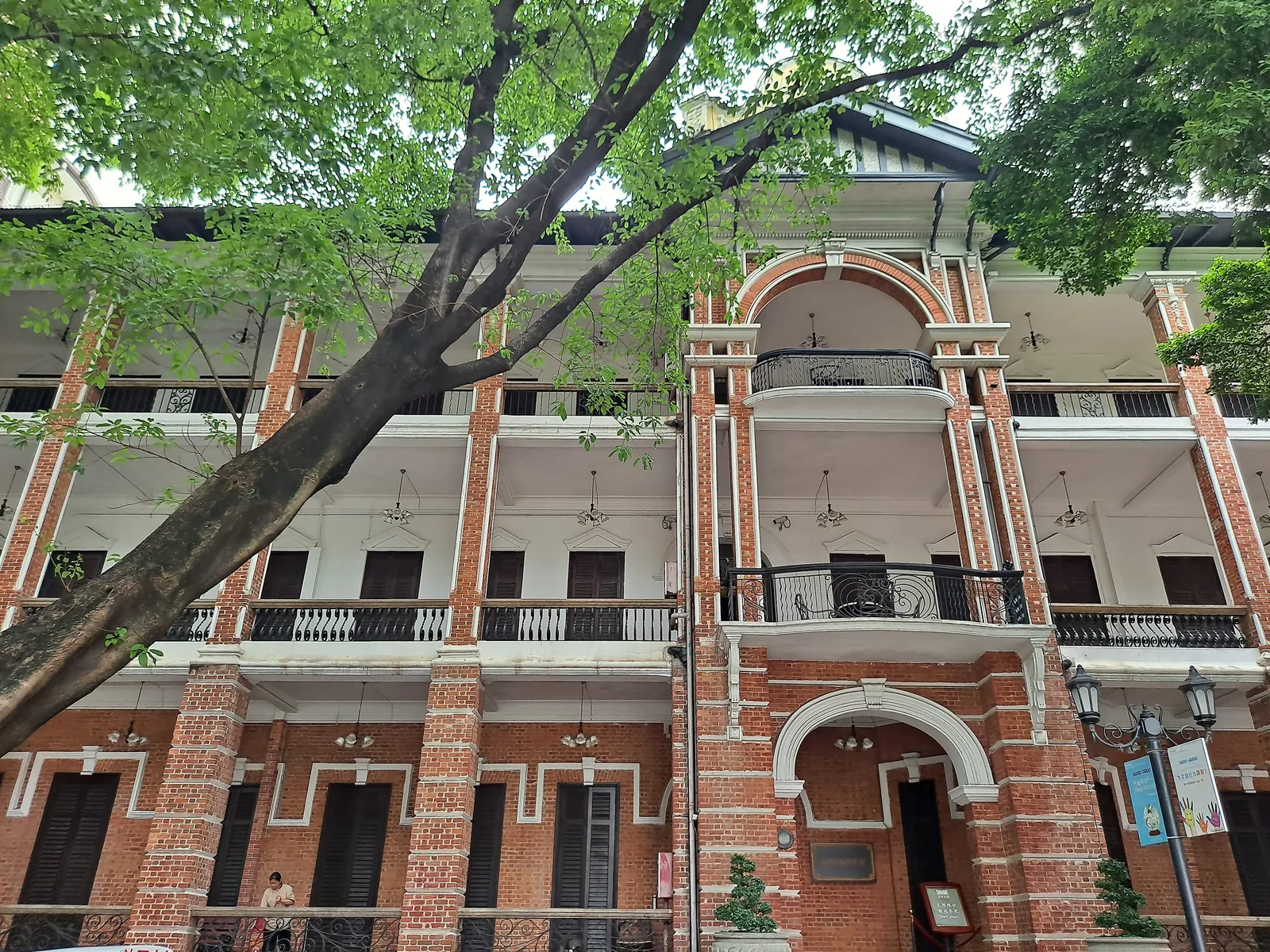 The red-brick front of the building today, banded in white, with arched balconies, behind a tree.
