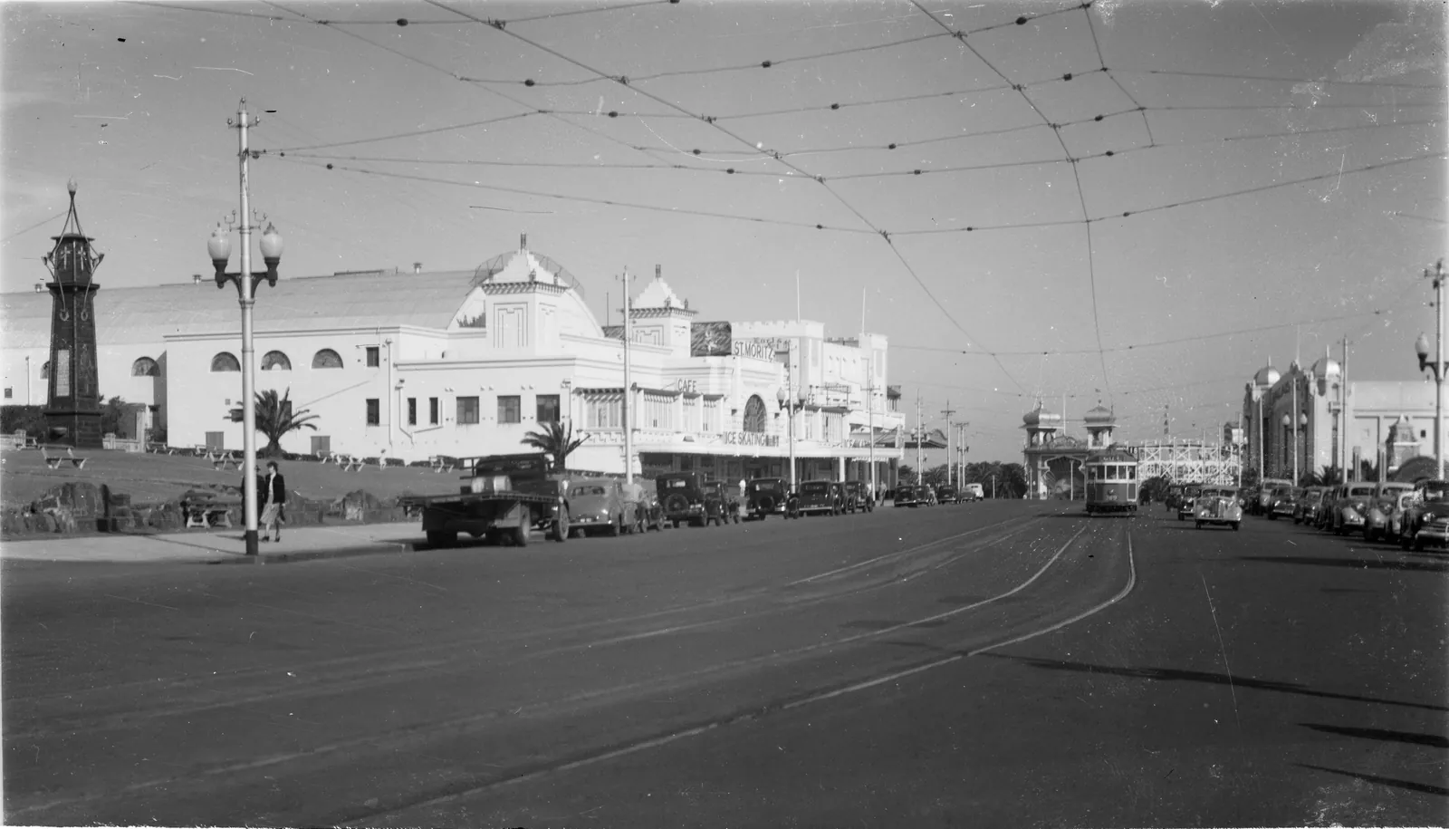 A wide road with tram tracks and overhead wires, a long white domed building on the left and cars parked along the kerb.